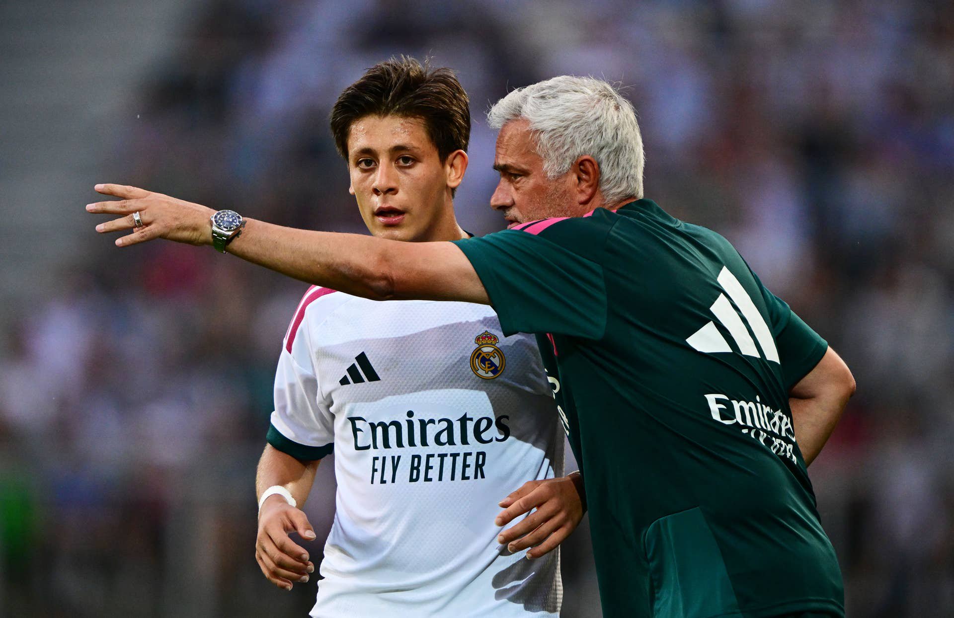 Arda Güler im weißen Real-Madrid-Trikot gestikuliert auf dem Spielfeld, während ihm ein Betreuer von hinten Anweisungen gibt.