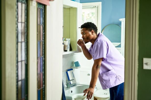 Black man in his 20s looking in mirror using toothbrush, leaning on edge of basin, getting ready for work in the morning