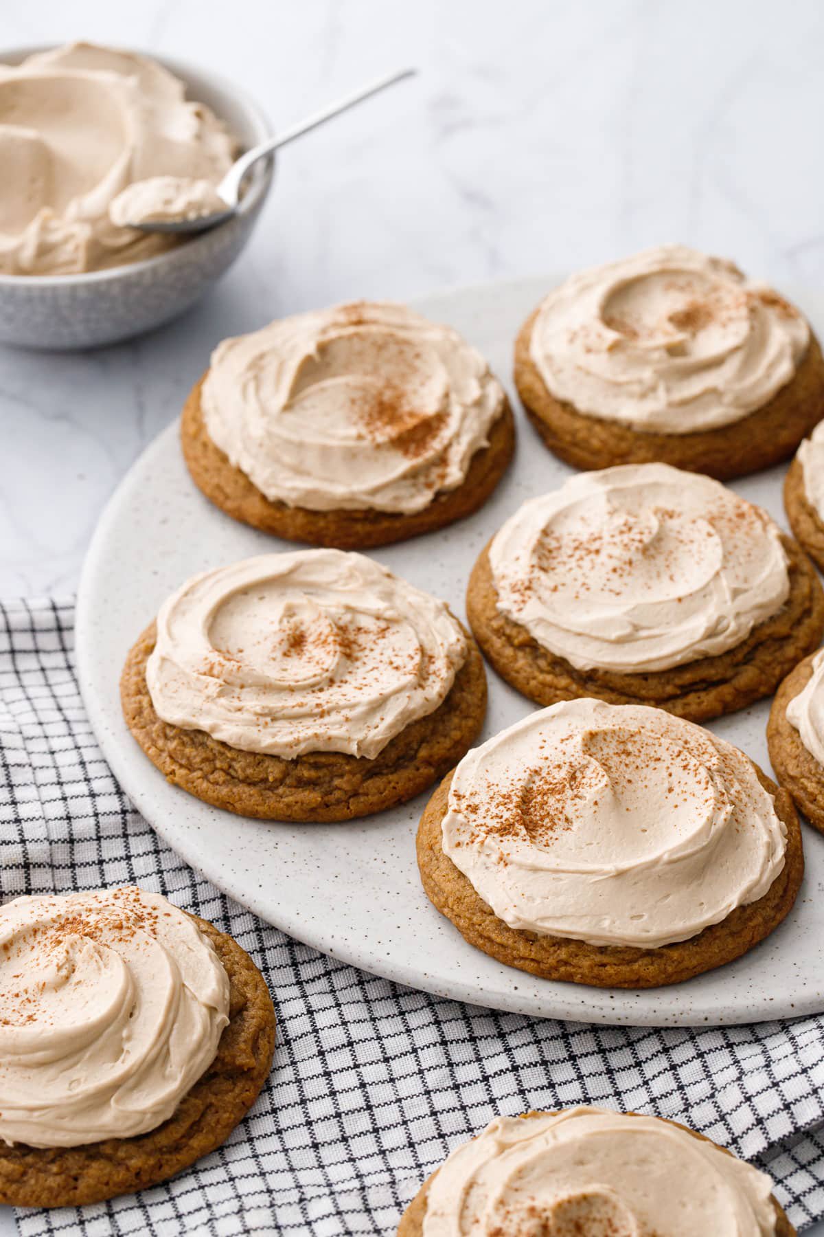 Round flat plate with Frosted Caramel Pumpkin Cookies dusted with cinnamon, more cookies on a striped napkin in the foreground and a bowl of frosting in the background.