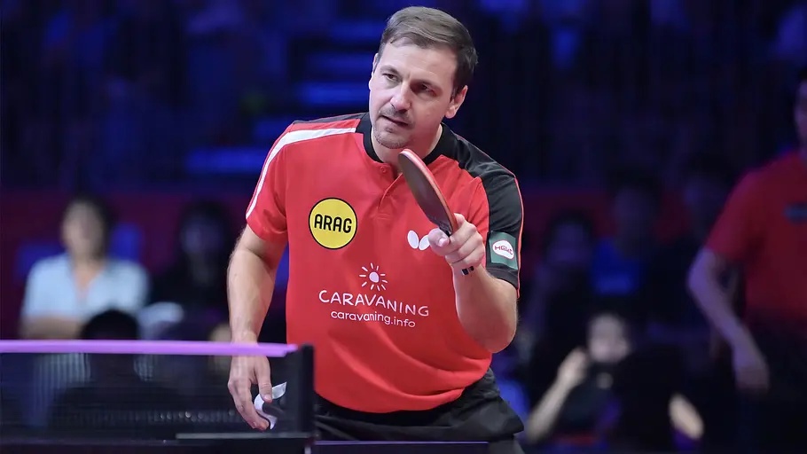 Timo Boll im roten Trikot hält seinen Schläger über dem Tisch. Im Hintergrund sitzen unscharf erkennbare Zuschauer.