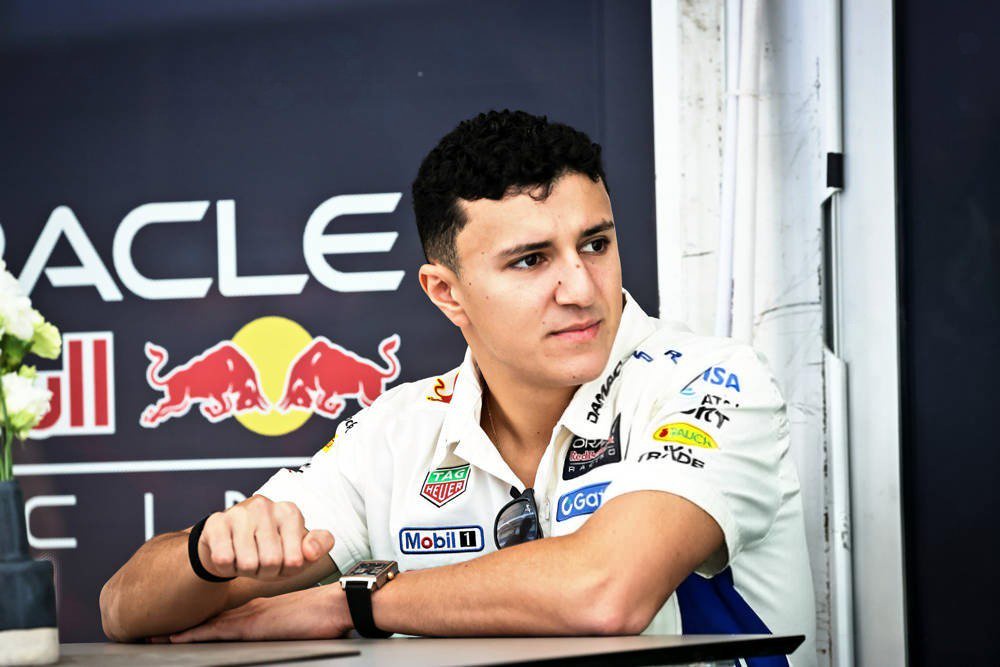 Racing driver in a white Red Bull-linked team shirt with Mobil 1 and Tag Heuer logos, seated at a table in front of a Red Bull Oracle backdrop.