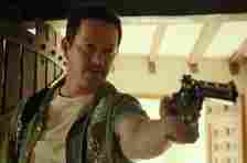 Person dressed casually in a T-shirt and open shirt, holding a revolver and aiming it forward. The setting appears to be indoors with wooden beams in the background