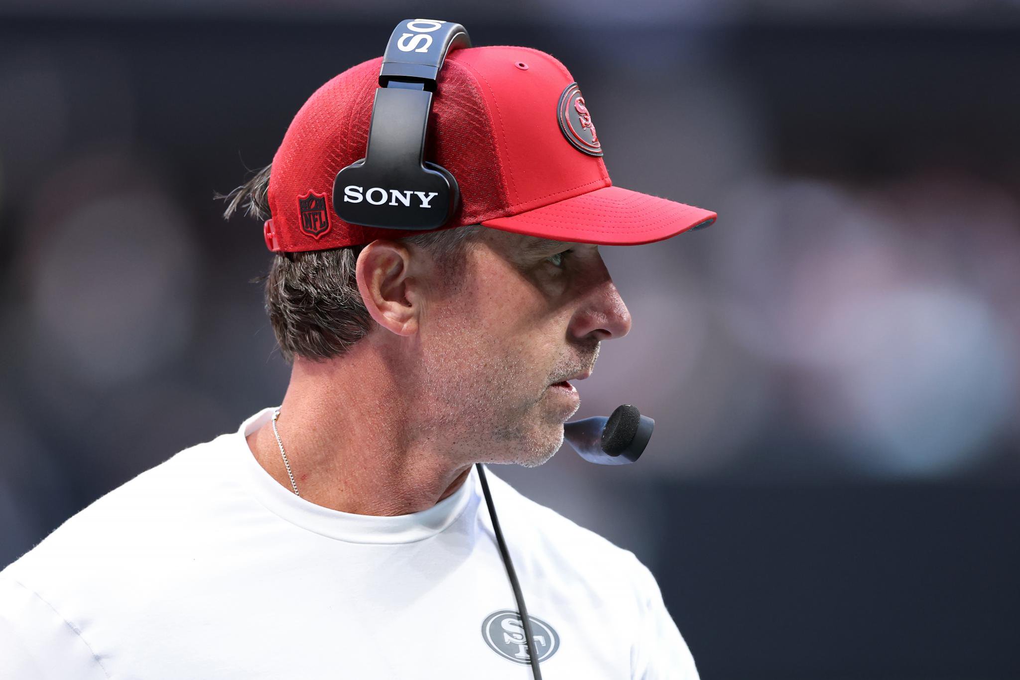 San Francisco 49ers coach Kyle Shanahan wearing a red cap and a headset.