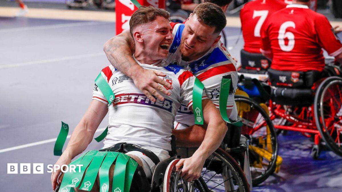 Rob Hawkins, seating in a wheelchair in a white kit with green tags on his shoulders and knees, is hugged from behind by