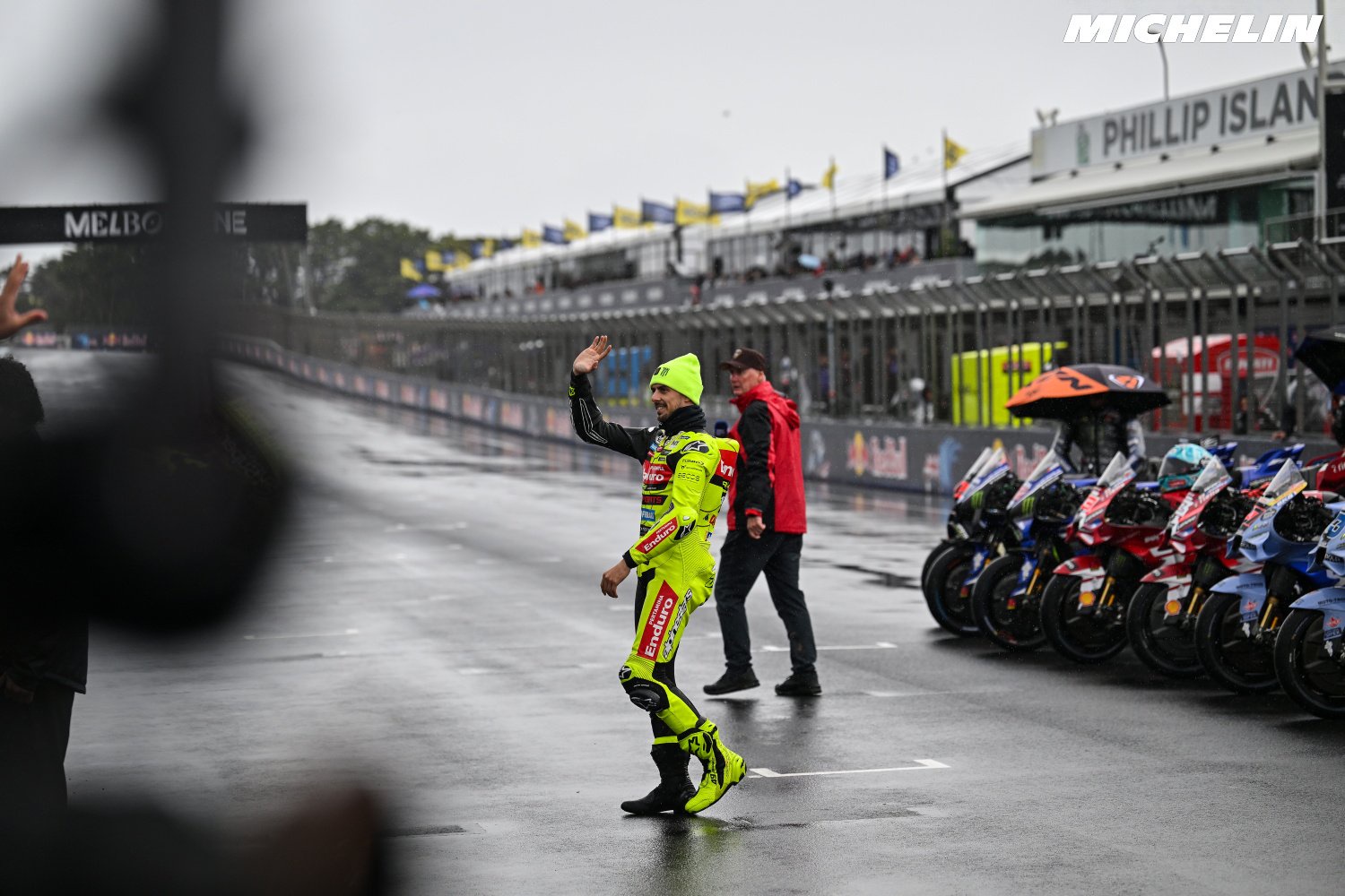 A rider in yellow-green leathers waves to fans on a wet MotoGP grid at Phillip Island, with rows of bikes behind him.