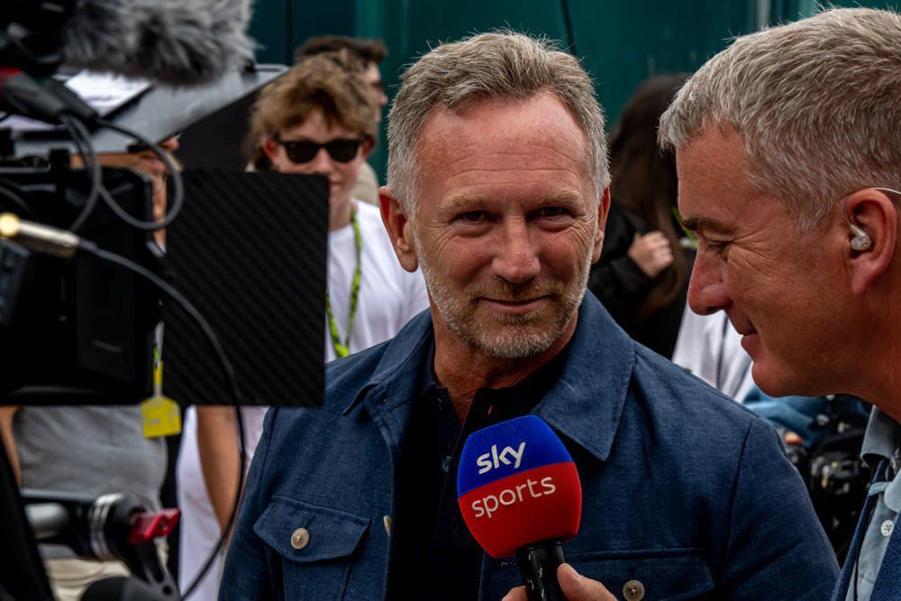Man with grey hair and stubble in a denim jacket speaking to a Sky Sports reporter holding a Sky Sports microphone in a paddock area