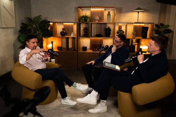 Three individuals are engaged in a discussion while seated on beige armchairs in an elegantly furnished room. The room is adorned with decorative shelves filled with various items, and warm lighting illuminates the scene. Each person is holding a microphone, suggesting a formal or recorded setting.
