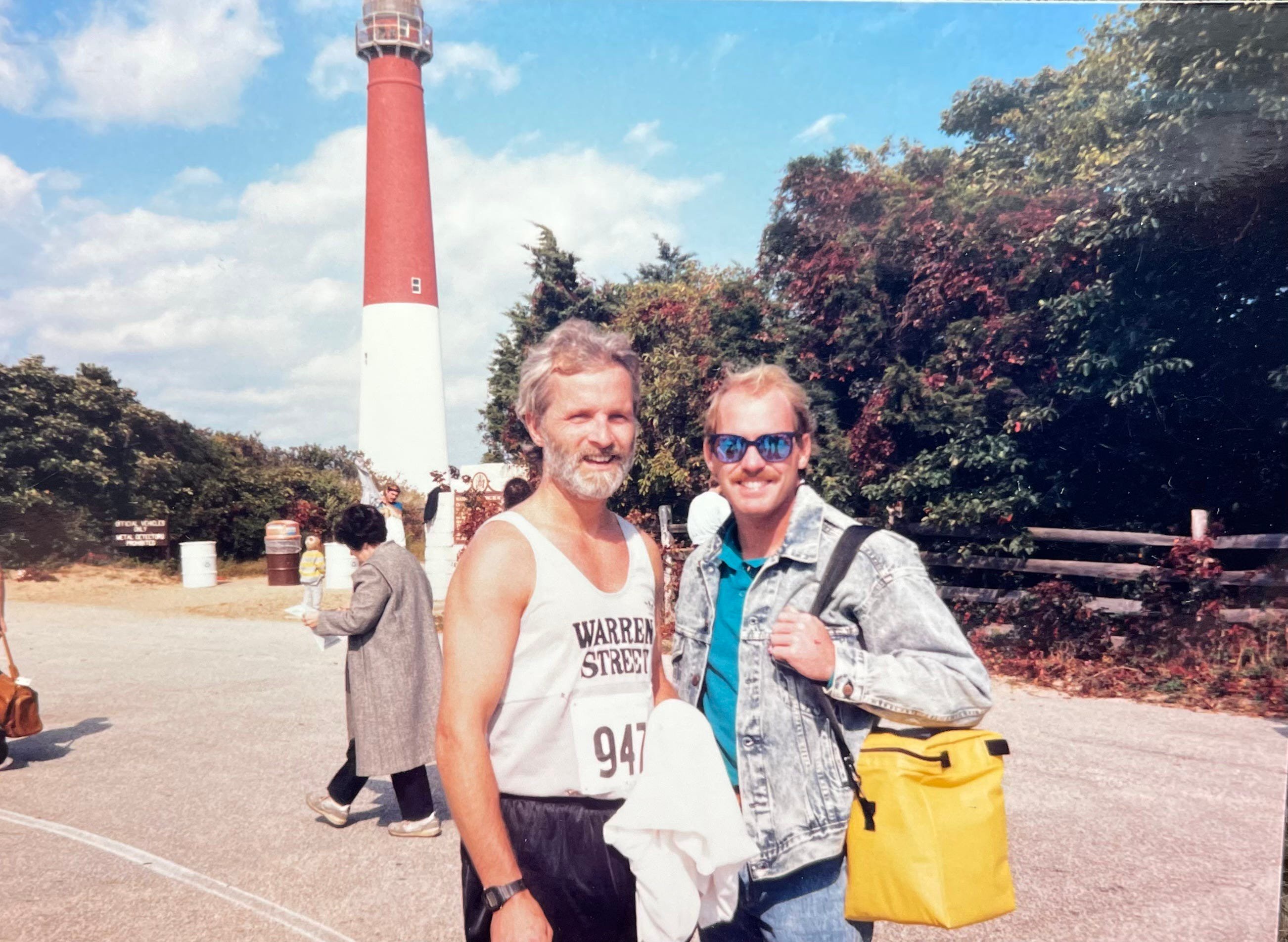 Two men posing together in front of a lighthouse on a sunny day, one wearing a tank top and the other in a denim jacket.