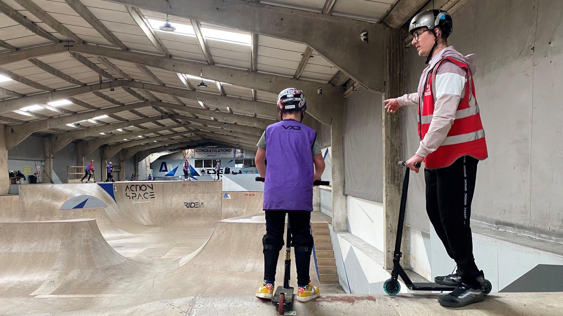 People scooting at an inside venue, with two people standing by a ramp. Jayden Sharman is to the right, wearing a red hi-vis jacket and black trousers. He has a helmet on. Other people are in the distance. There are ramps in the room and a large metal roof with lights in it.
