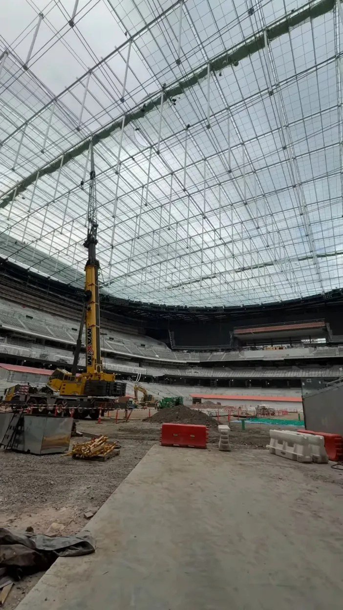 A large crane stands inside a large, dome-shaped stadium under construction, with a massive steel and glass roof structure overhead.