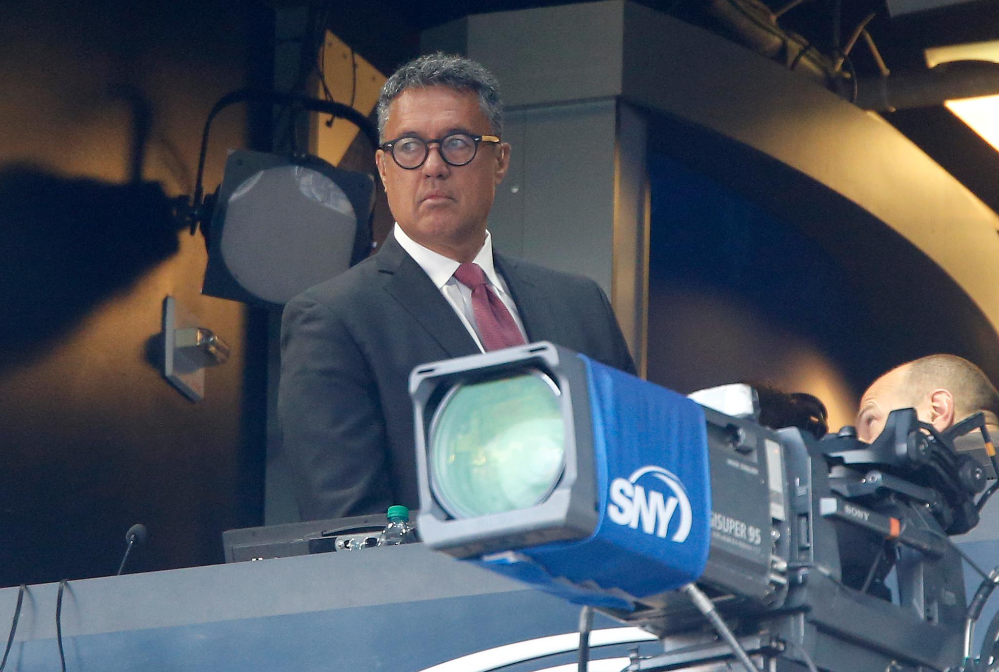 A man wearing glasses, a suit, and a tie stands behind a television camera with an SNY logo in a broadcast booth.