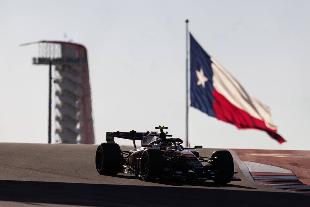 An F1 car with a dark livery and yellow helmet-visible accents on track near a Texas flag and grandstand at COTA, US Grand Prix