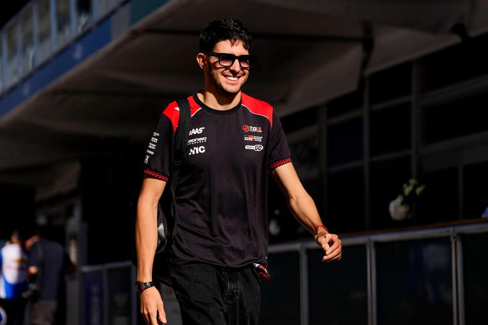 A smiling man in sunglasses and a black Haas F1 team shirt walks through a paddock area, carrying a backpack.
