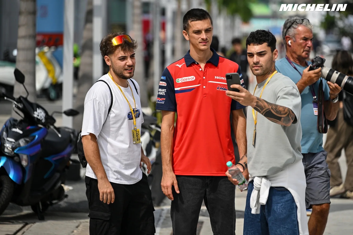Three young men on a street, one in a red Castrol Honda team shirt checking a phone, with a photographer behind; MotoGP paddock scene