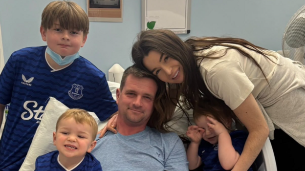 A family of five - three young boys, a man and a woman gather around the man in a hospital bed.