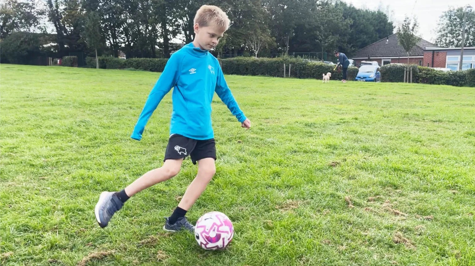 A boy in a blue training top and black shorts is playing football in a park field.