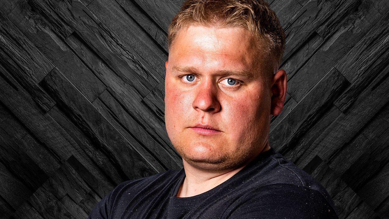 A person with short hair stands in front of a dark patterned wooden backdrop. The person is wearing a black T-shirt and is facing the camera.