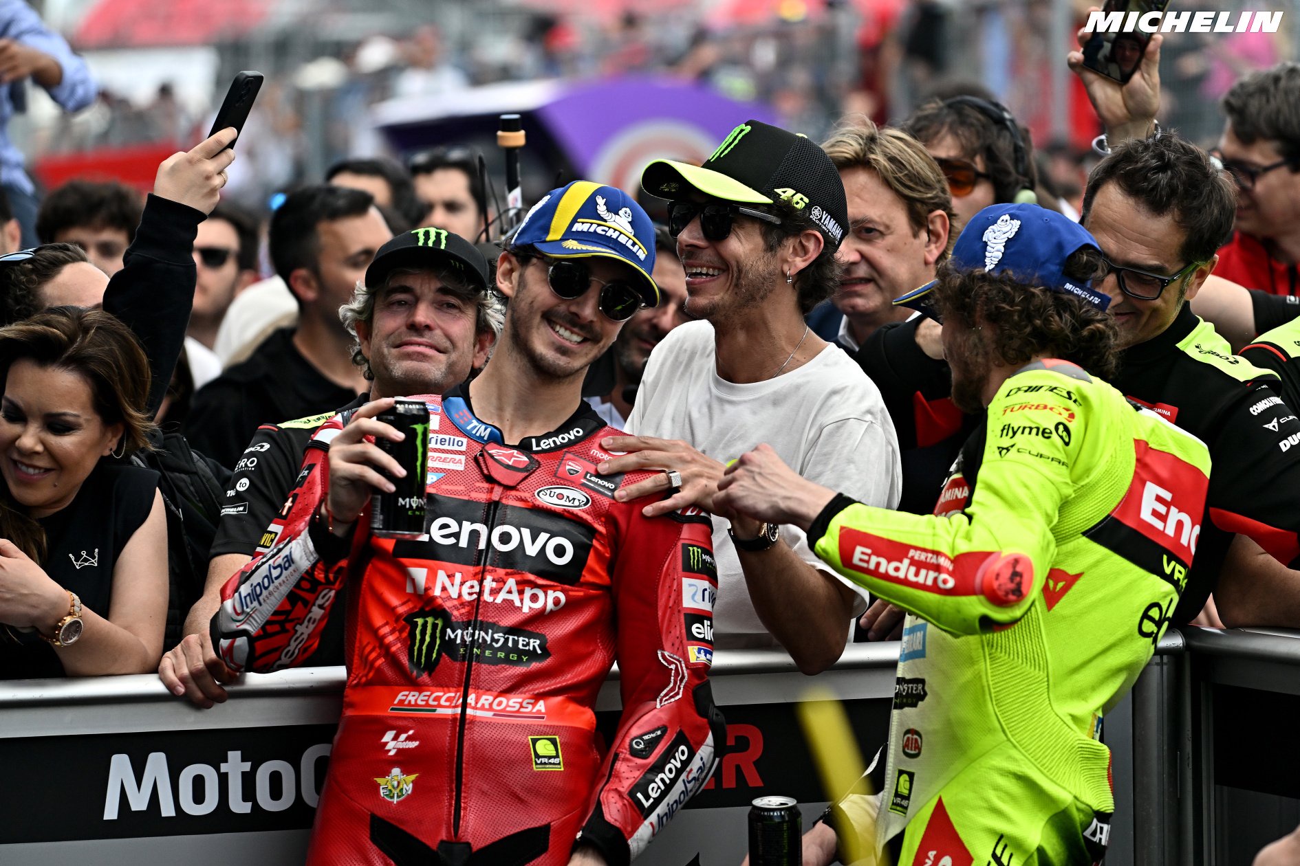 Rider in red Ducati leathers with Lenovo and NetApp logos poses for a selfie with fans at a MotoGP event, Michelin branding visible.