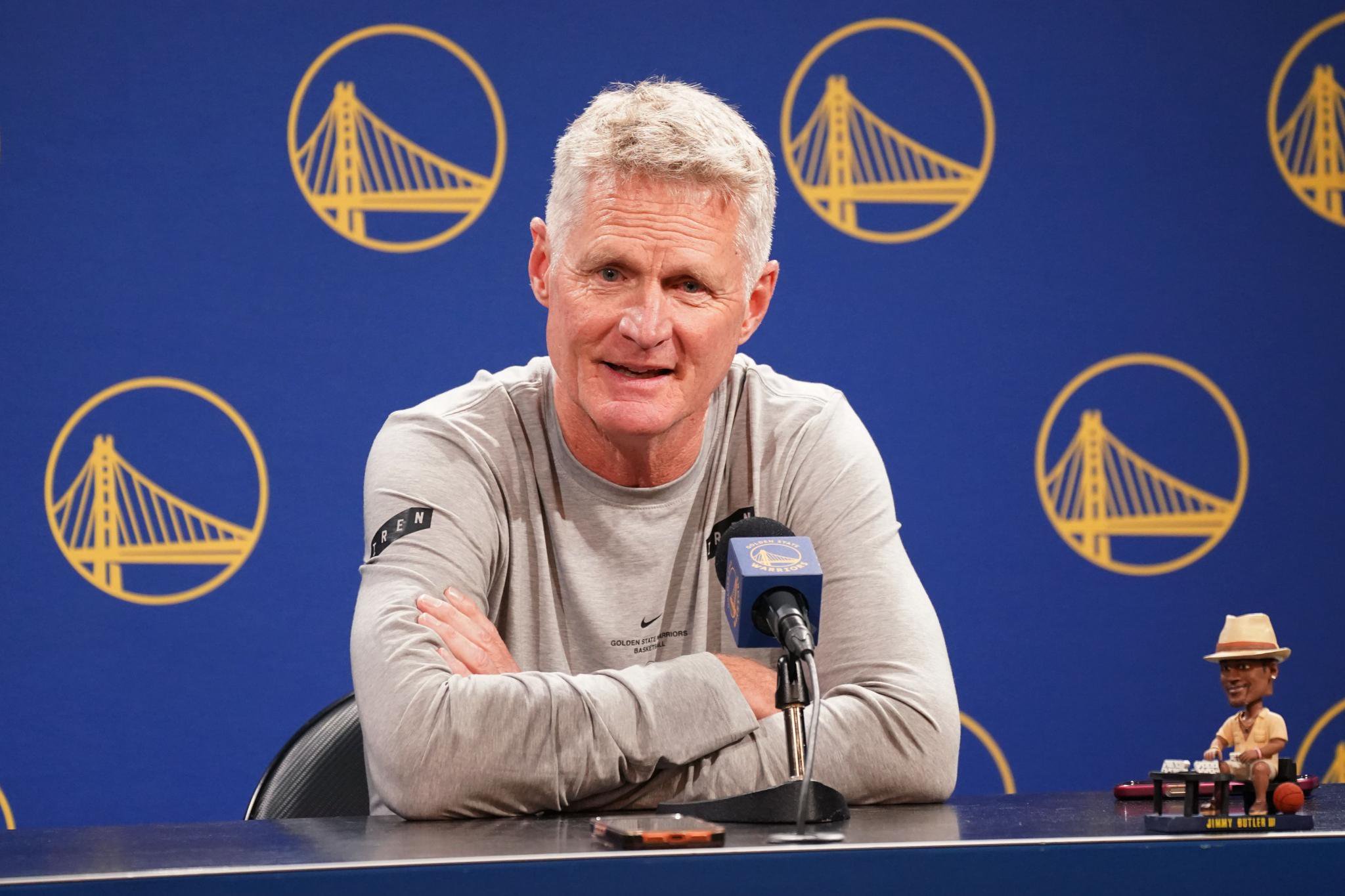 Golden State Warriors head coach Steve Kerr speaks at a press conference in front of a blue backdrop with team logos.