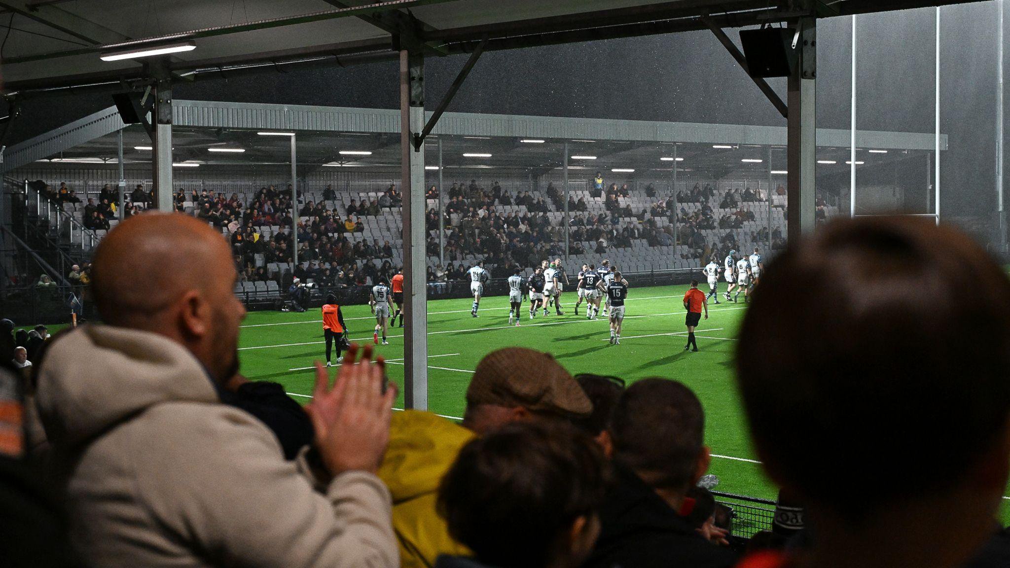 Fans in the foreground applaud players on the pitch in front of half-full stand