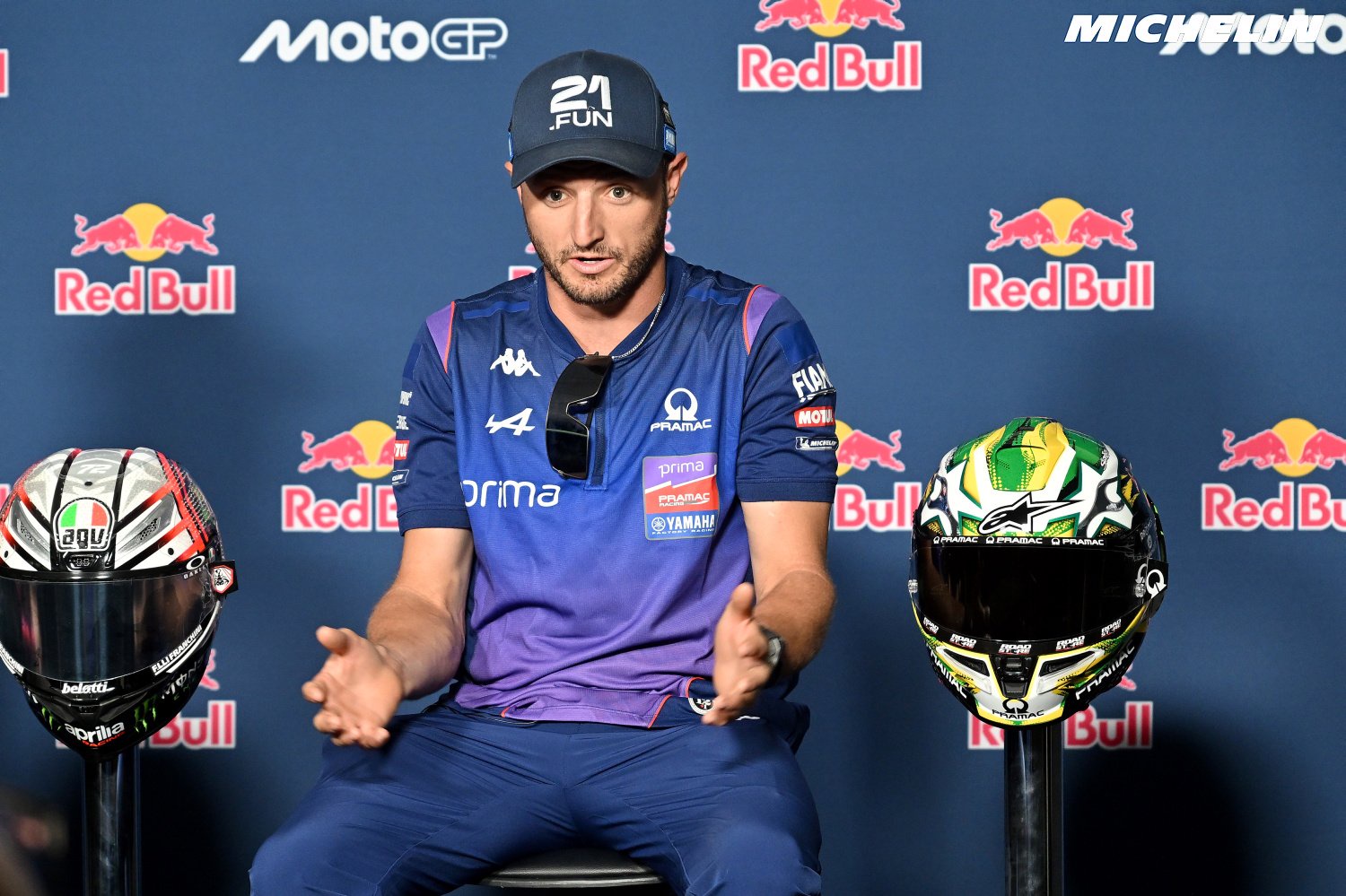 Rider in a blue Prima Pramac Yamaha shirt and cap seated at a MotoGP press conference, gesturing, with two helmets displayed beside him.