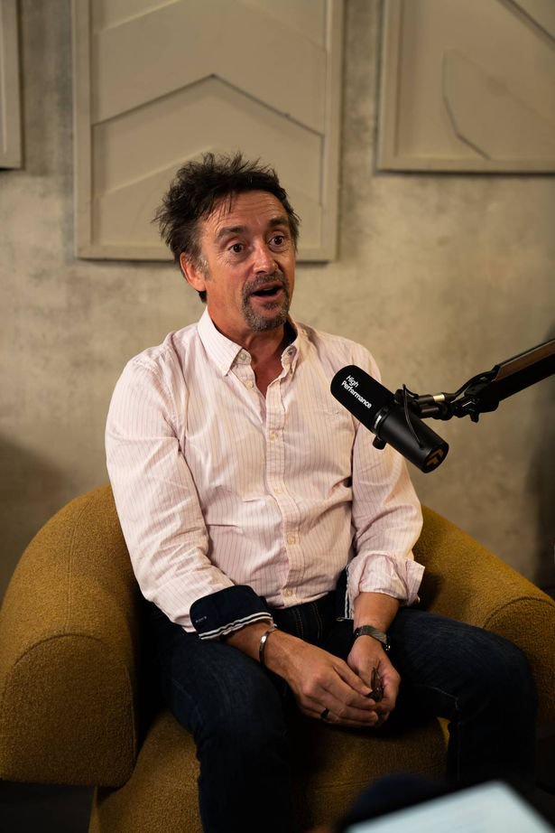 An individual is engaged in an interview, addressing the media with a focused expression while standing before a microphone. The background features a textured wall with a decorative frame.