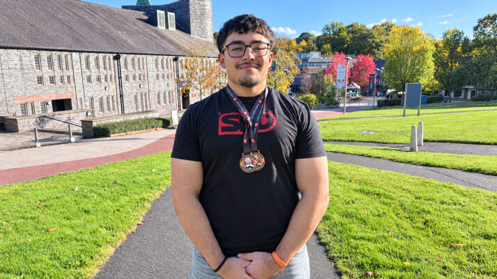 A man stands outdoors wearing competition medals around his neck, with campus buildings and green space in the background.