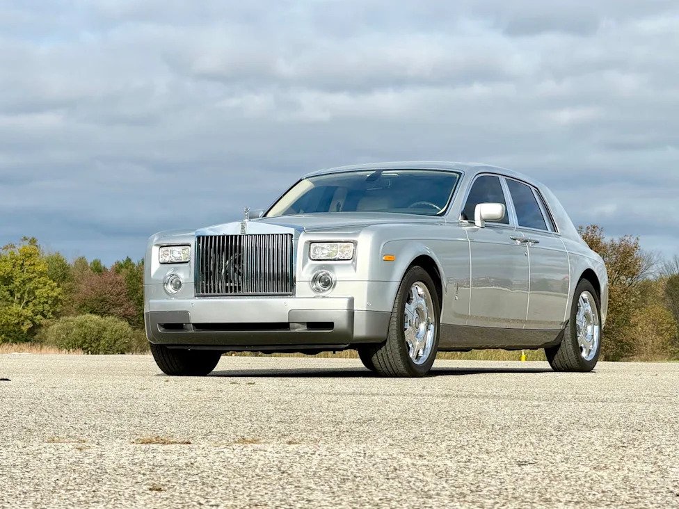 front three-quarter view, silver luxury sedan parked on pavement with trees and cloudy sky.
