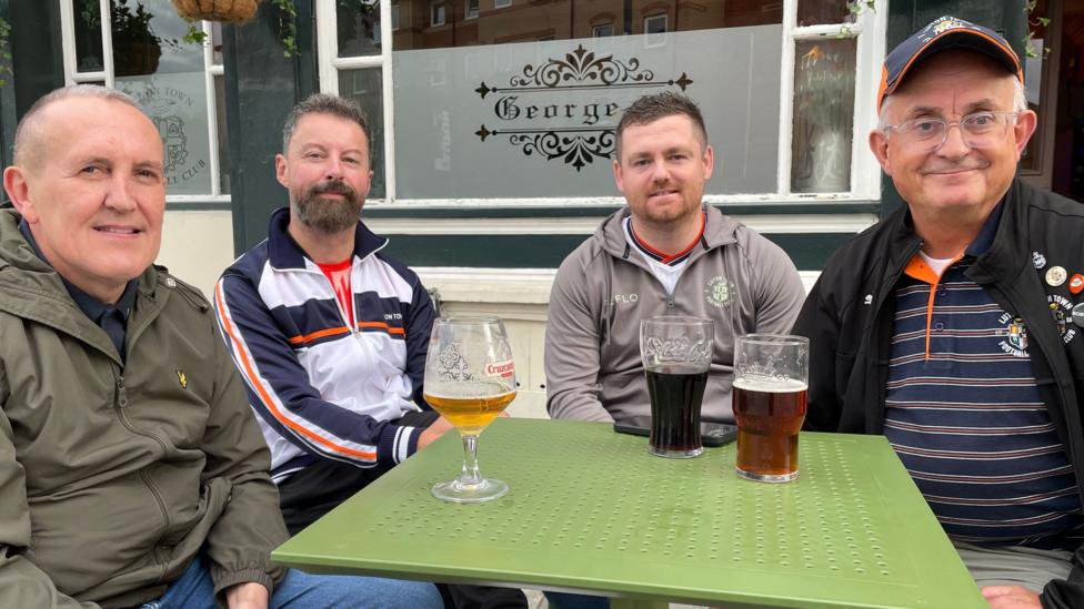 Four Luton fans are sitting around a green table with a mixture of drinks  outside a local Luton pub. 
