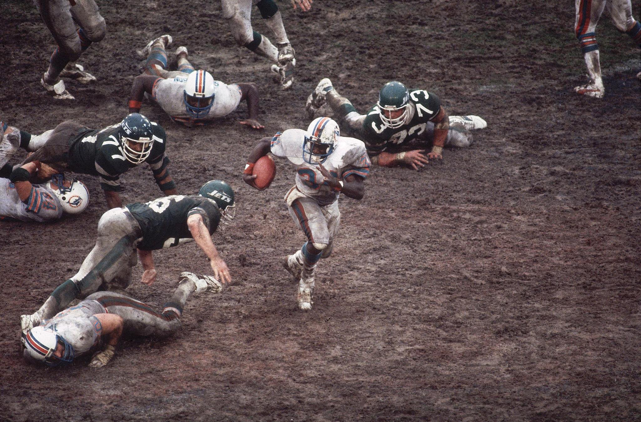 A Miami Dolphins player runs with the football on a muddy field during the 1983 AFC Championship game against the New York Jets, surrounded by fallen players.