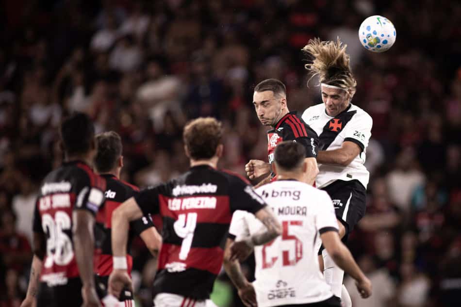 Flamengo x Vasco; jogador rubro-negro salta para cabecear a bola, cercado por adversários, torcida ao fundo.