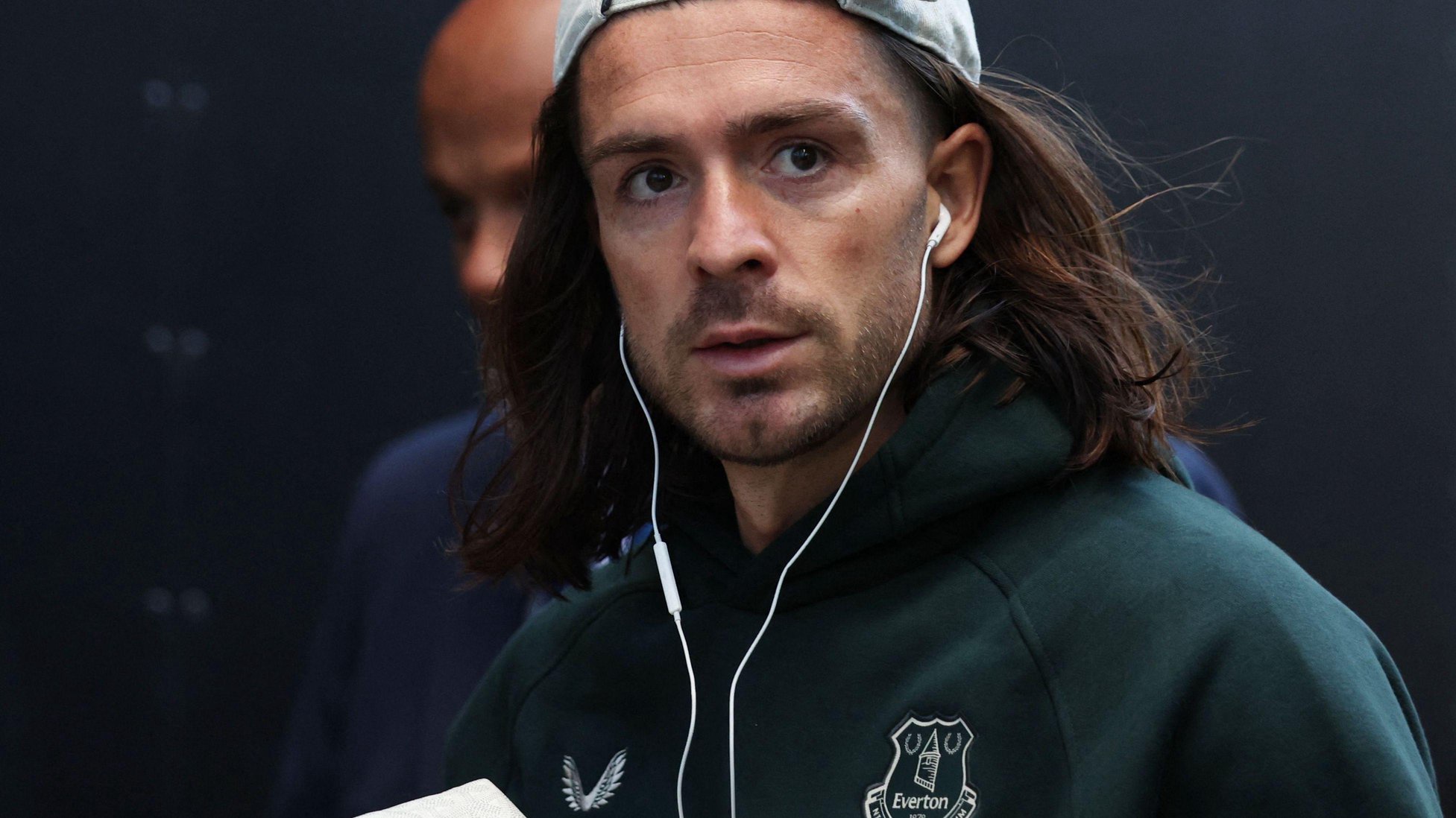 A man  wears a cap and has dark flowing long hair. He wears wired headphone and a tracksuit top with the Everton badge on it. 