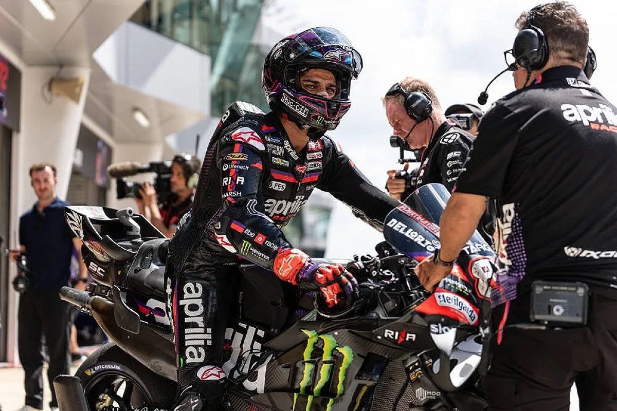 Rider in Aprilia leathers and helmet beside his Aprilia MotoGP bike in the pit lane, with team crew in black Aprilia shirts