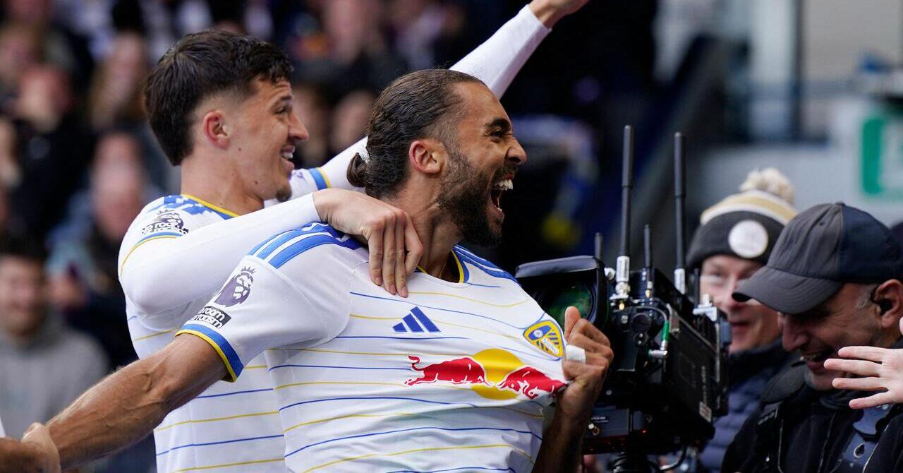 Footballer Dominic Calvert-Lewin looking to his left in a stadium and smiling and shouting to celebrate a goal while playing for Premier League team Leeds United in a white kit with blue trim and yellow stripes. Calvert Lewin is near two camera operators and being embraced by a teammate behind him with the Red Bull logo in red and yellow, the Adidas logo in blue and the club badge on his chest.