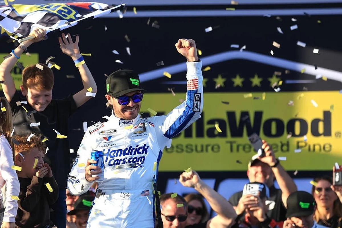 Driver in white Hendrick Cars.com suit and cap celebrating on stage with raised fist amid confetti, NASCAR Cup victory celebration