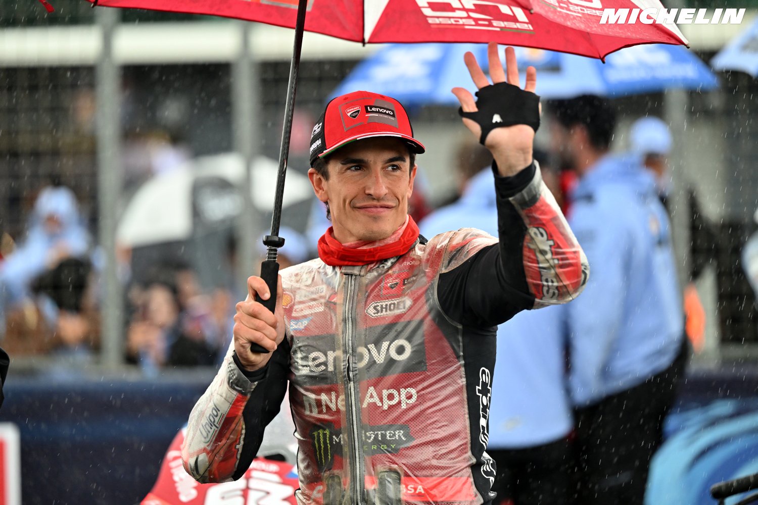 A rider in a red Ducati-team cap and wet red-and-white leathers waves under a red umbrella in the rain at a MotoGP grid, with a Michelin logo in the corner