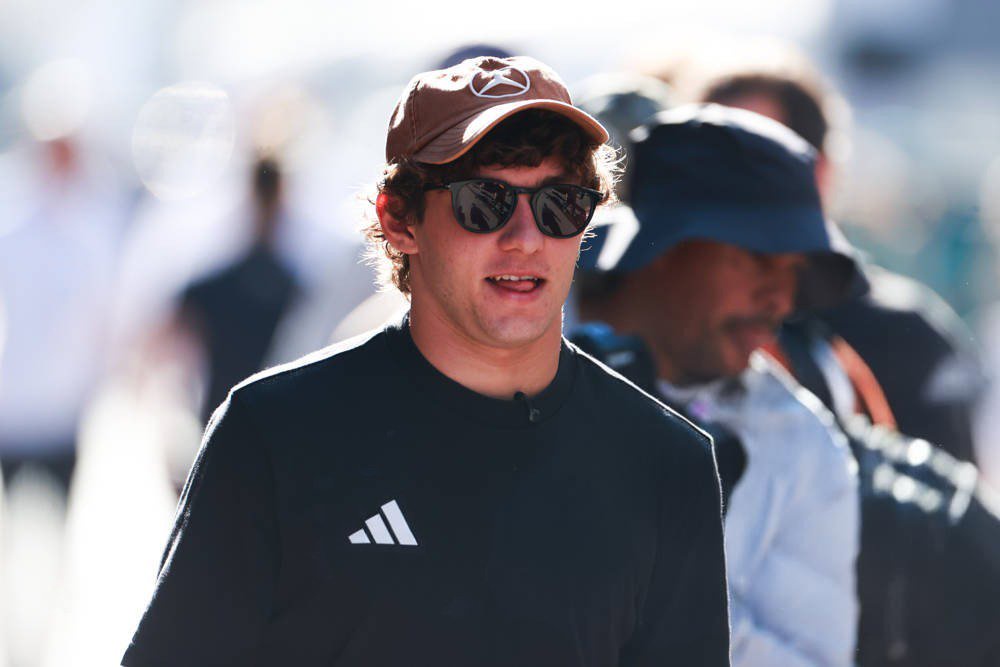 Driver in a black Adidas sweatshirt, brown cap and sunglasses in a blurred paddock crowd, consistent with the Formula 1 article on Andrea Kimi Antonelli