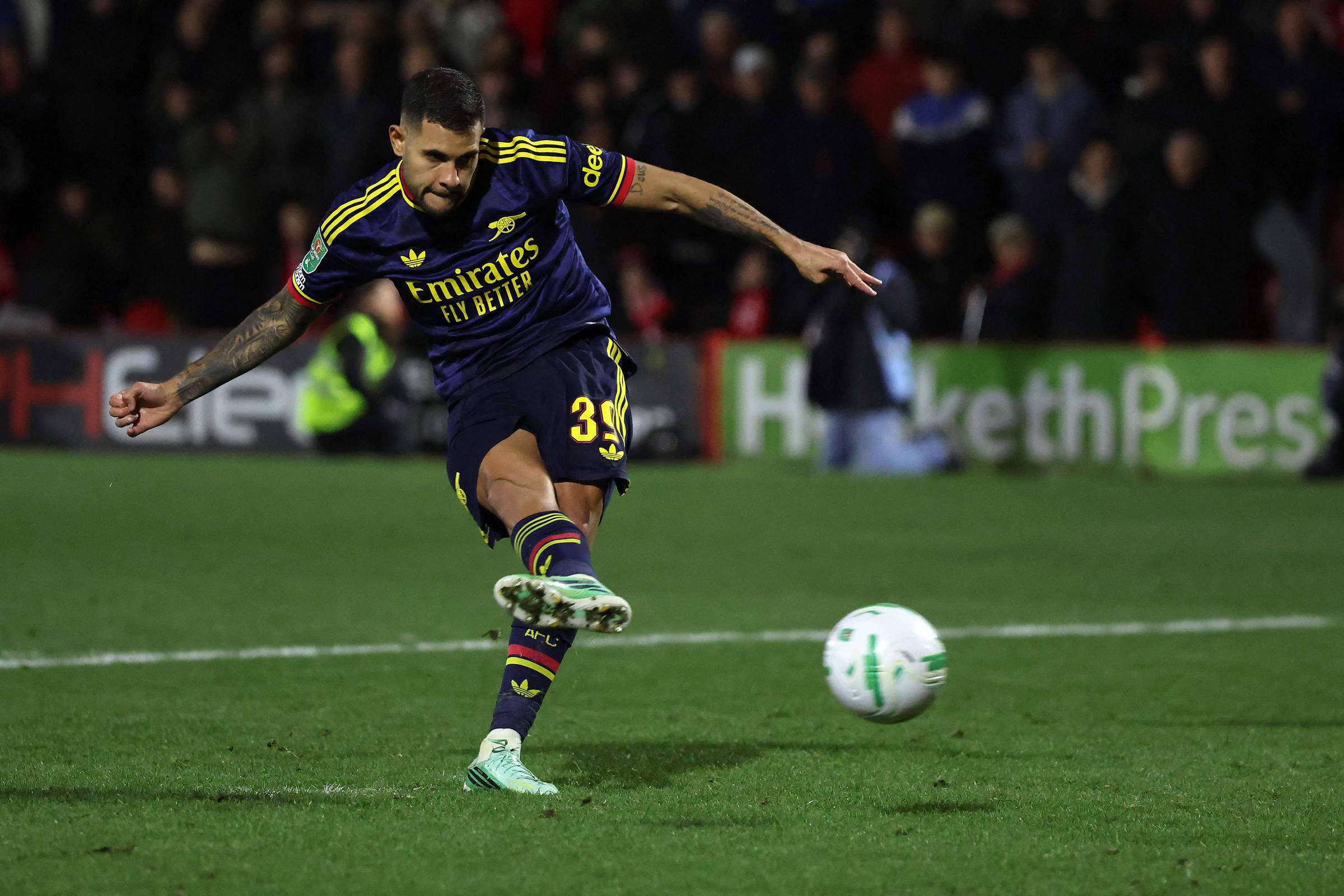 Jogador do Arsenal com uniforme azul escuro e detalhes amarelos chuta a bola durante jogo noturno em campo gramado. Ele é Bruno Guimarães. Público desfocado ao fundo observa a ação.