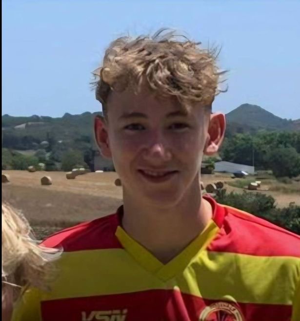 An individual stands outdoors, smiling broadly, against a backdrop of rolling hills and greenery. The person is dressed in a vibrant red and yellow sports jersey. The sky is clear and blue, indicating a sunny day.
