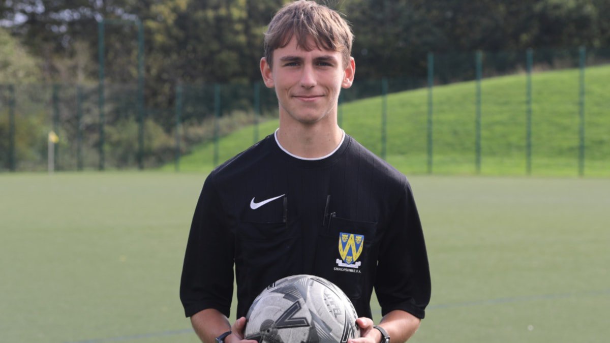 A young man with short blonde hair in a black referees uniform holding a white football on a football pitch