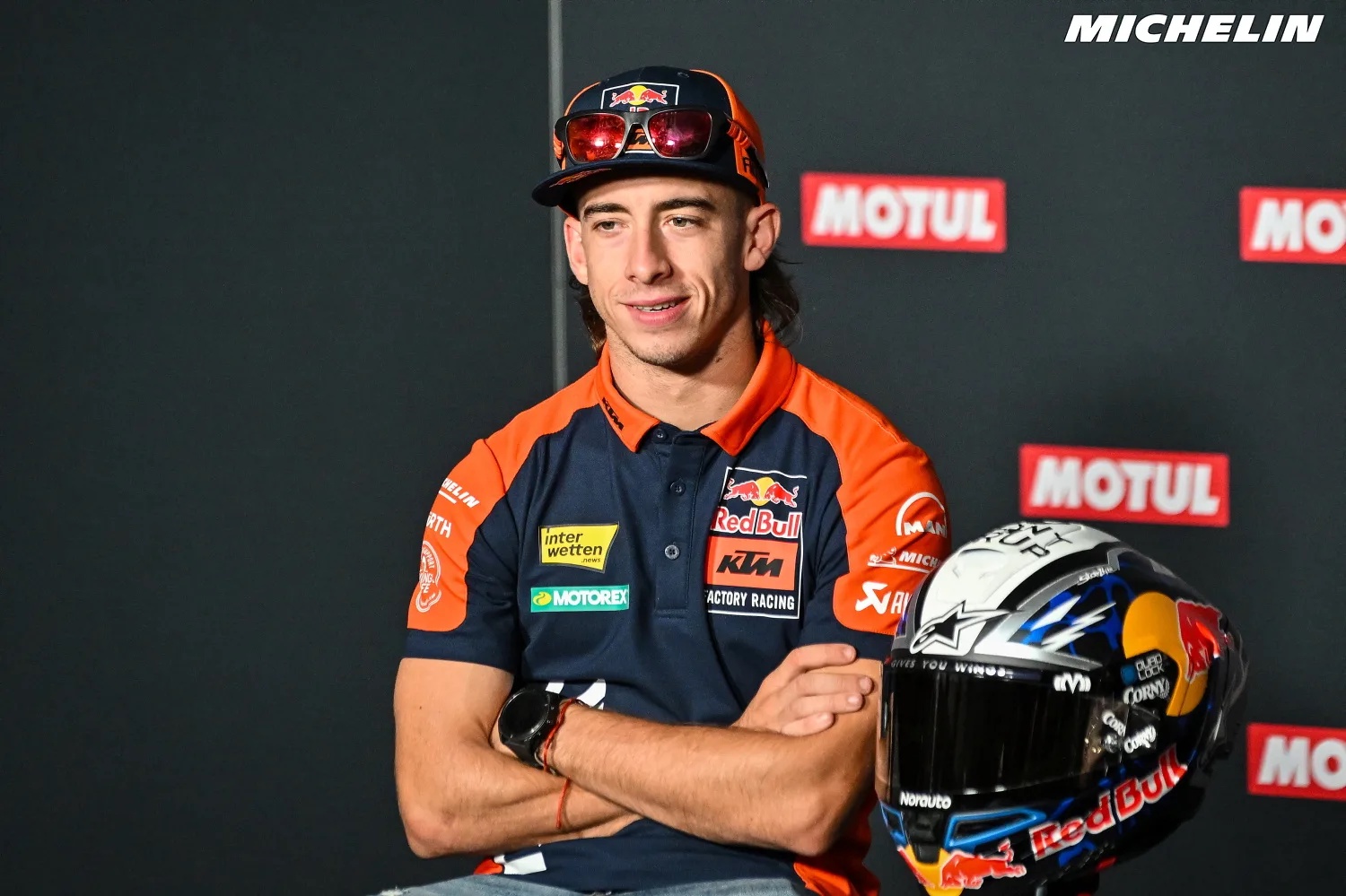 Rider in KTM Factory Racing polo and cap with a Red Bull helmet, arms crossed at a MotoGP press conference, Motul backdrop