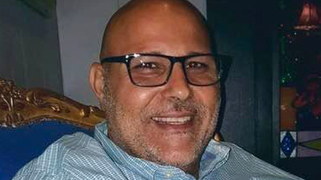 A close-up portrait of a bald, middle-aged man with glasses and stubble, wearing a light blue checkered shirt.