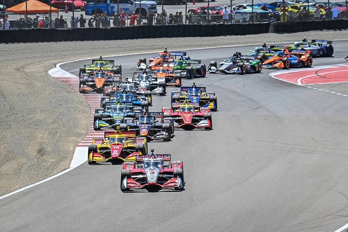 IndyCar cars racing in a pack through a turn on a road course, with a crowd watching from the fence line