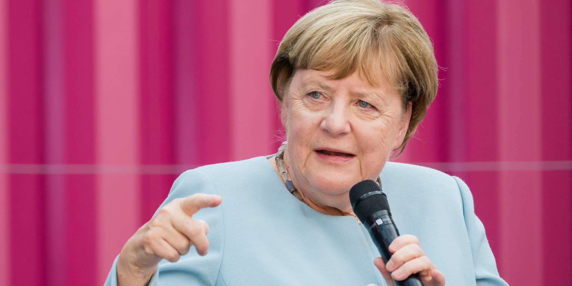 Frau in blauem Blazer spricht mit Mikrofon vor einem pinken gestreiften Hintergrund.