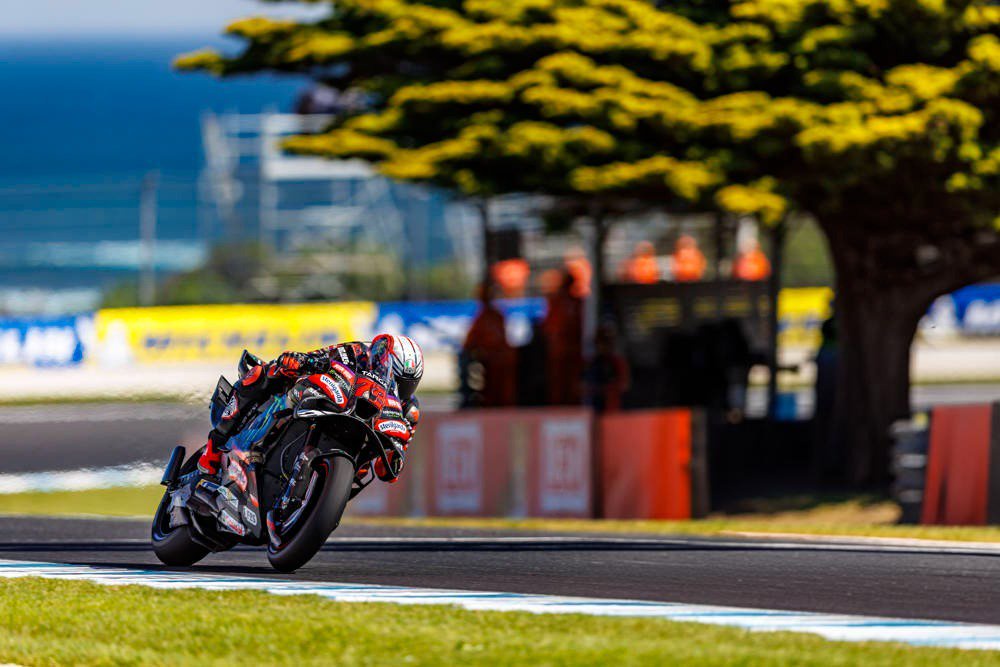 Aprilia MotoGP rider in red and black leathers leaning into a corner at the Australian Grand Prix circuit, with ocean behind