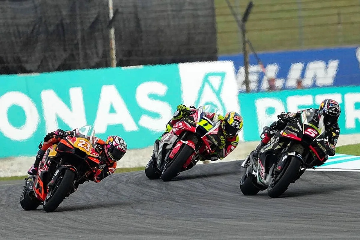 MotoGP riders racing at Sepang, with rider #23 in orange-and-black leathers leading two others around a corner past Petronas and Michelin signage
