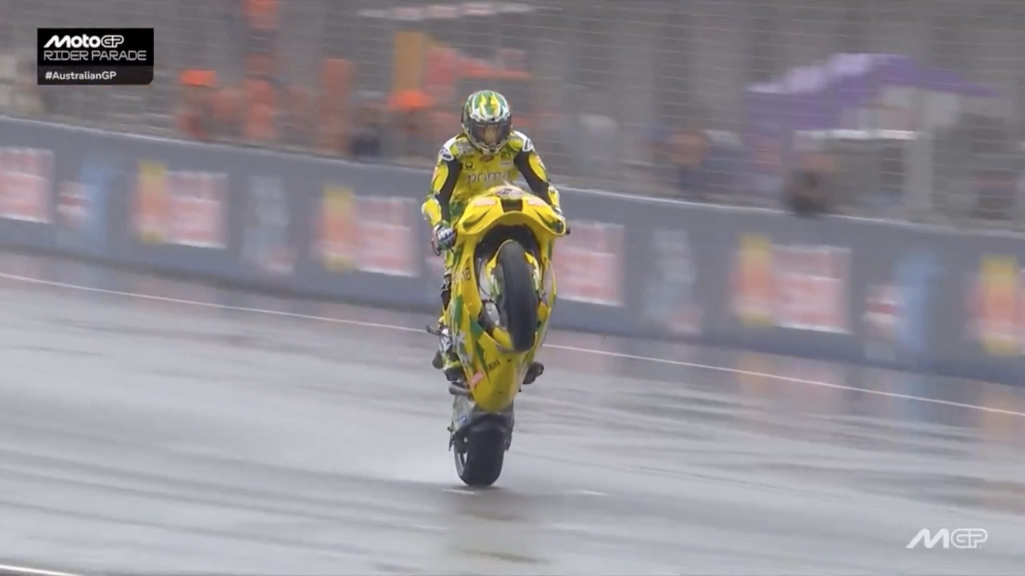 Rider in yellow and black leathers performs a wheelie on a yellow MotoGP bike on a wet track during the Australian GP rider parade