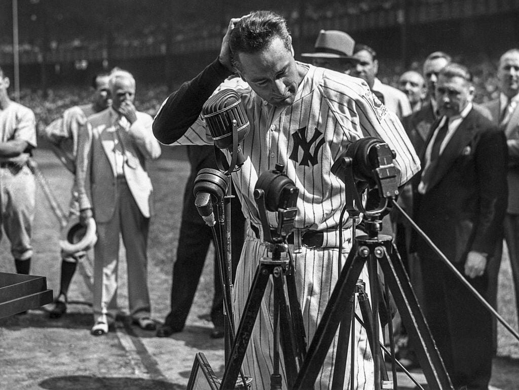 Baseball player in a New York Yankees uniform stands at a podium with multiple microphones, addressing a crowd on a baseball field. He looks down, touching his head, while people watch in the background. The setting appears historic.
