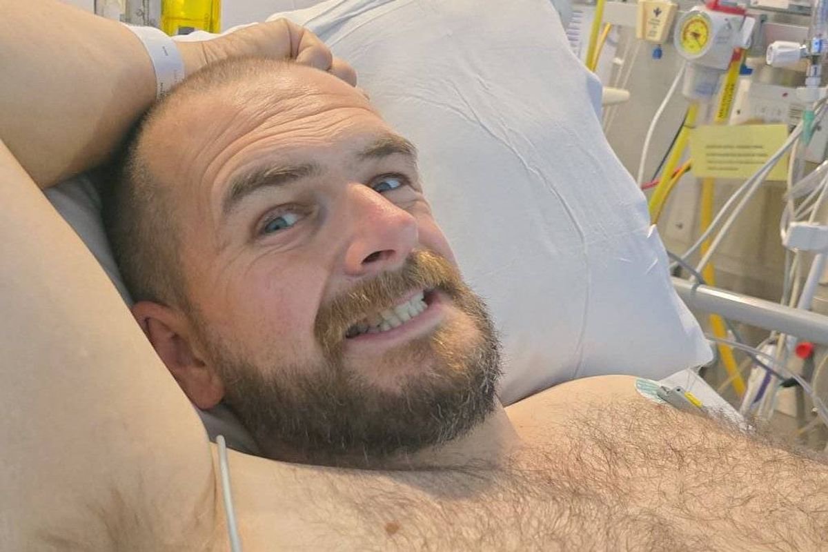 An individual, with a beard and short hair, is lying in a hospital bed, appearing to be in a state of recovery or rest. The person is wearing a hospital gown and has medical equipment visible in the background, indicating the presence of healthcare monitoring. The individual's expression is one of contentment or relief, suggesting a positive outlook or recovery progress.