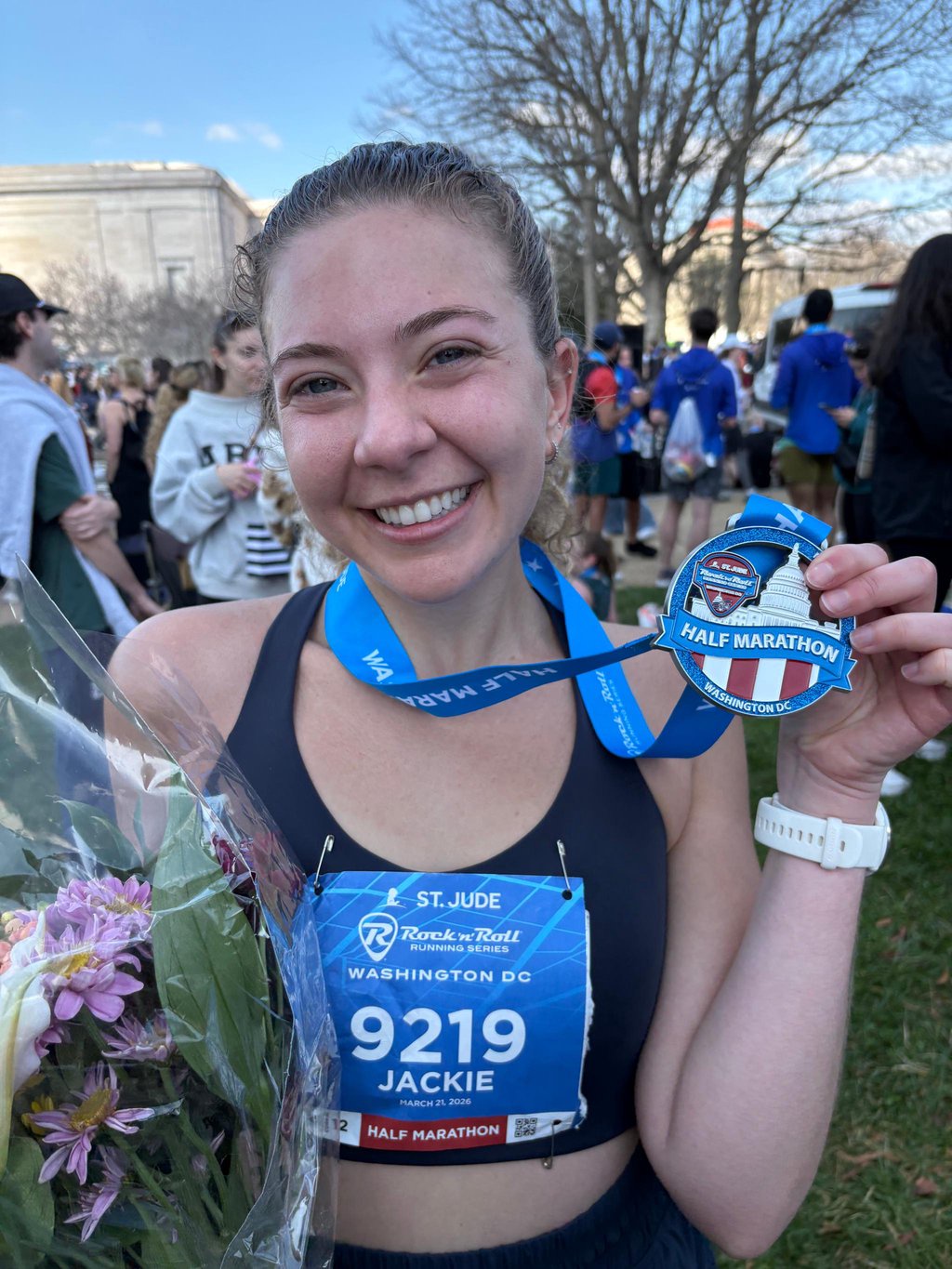 Jaclyn Corin smiles while holding a half marathon medal and a bouquet of flowers.